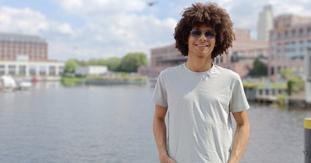 Jhorman steht vor einem Wasserbecken, dahinter Häuser, er lächelt in die Kamera